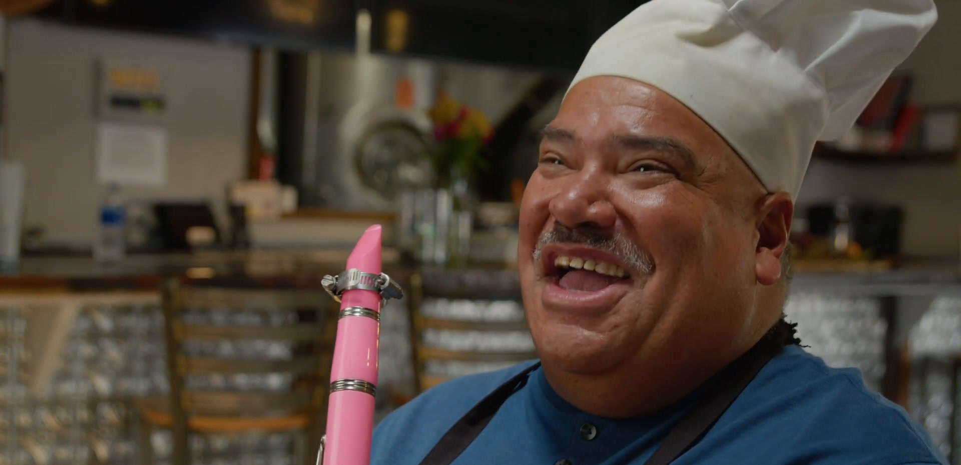 Jorge in a chef's hat and apron at the Brewability bar, holding up a pink clarinet and grinning.