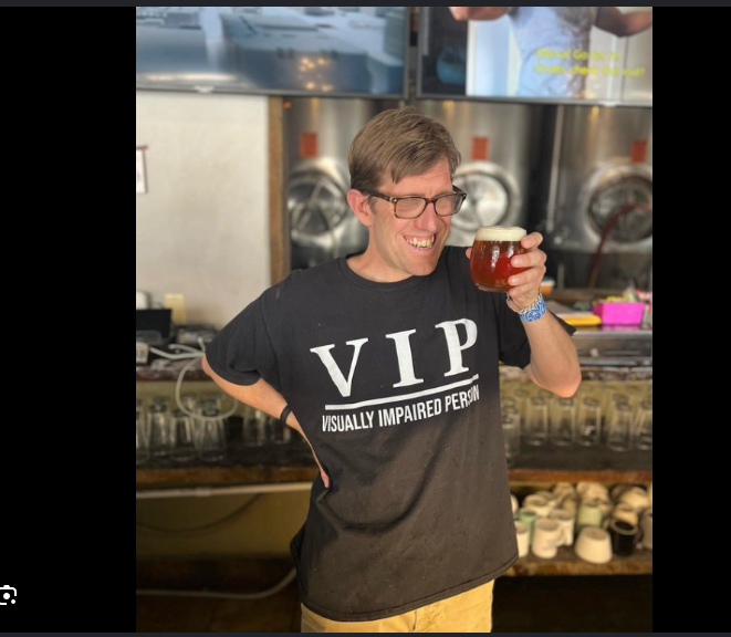 A Brewability team member smiling and holding a glass of amber beer in front of the colour-coded tap wall.