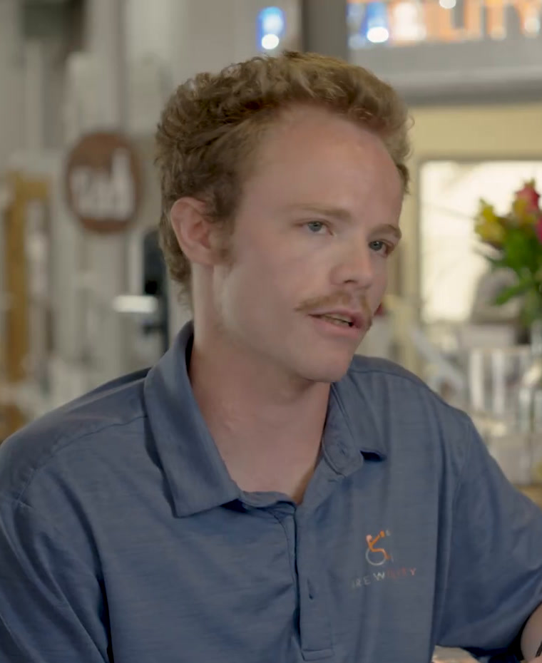 Avery in a blue Brewability polo shirt, sitting at the bar in front of a row of flowers, mid-conversation.