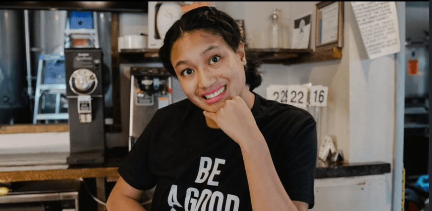 Miranda smiling beside the coffee equipment at Brewability, wearing a black Be a Good Person shirt.