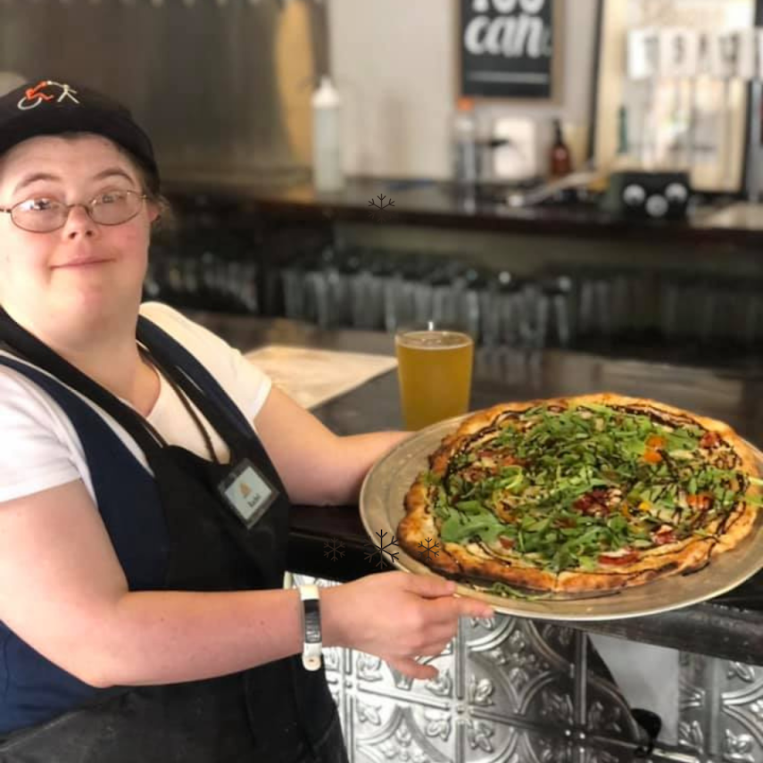 Rachel at the Brewability bar in her apron and name badge, holding out a large arugula and balsamic pizza she is about to serve.